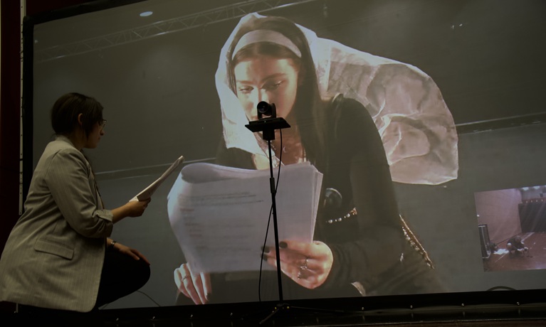 A female student holding a script next to a large screen showing another student elsewhere also reading from a script