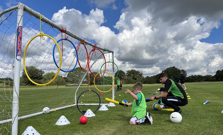 A boy holding a foam bat kneeling down on grass alongside a Coventry University researcher next to a football goal with coloured hoops attached to the crossbar