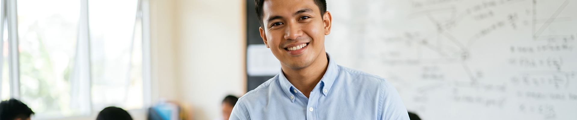 A young male teacher stood in a classroom holding a whiteboard marker