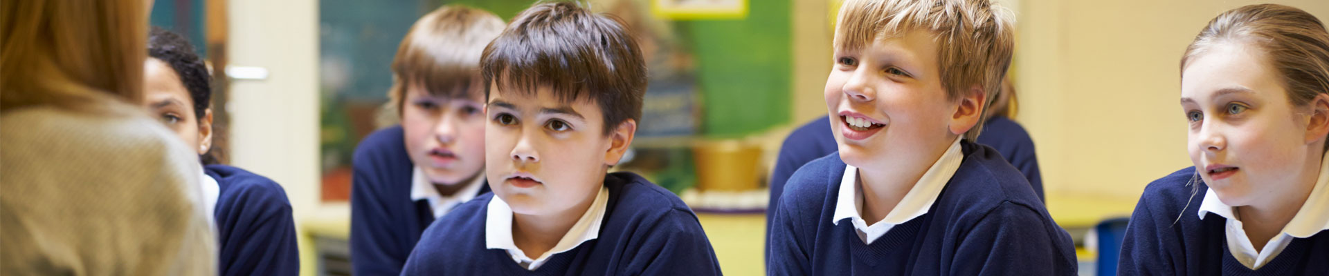 Primary schoolchildren sat at desks as a teacher talks to them from the front of the classroom