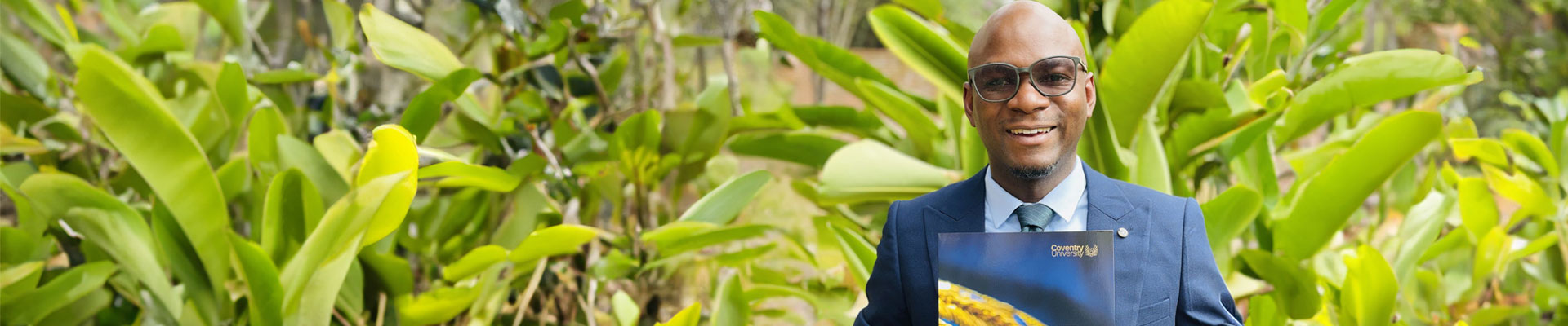 Chilipo Gunda stood outside against a green jungle plants background, holding his MA in Educational Leadership certificate from Coventry University