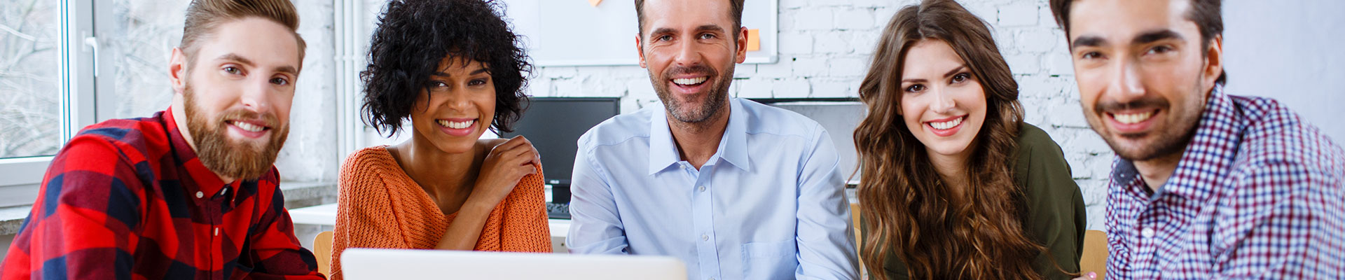 A group of people of various ages sat at a table with a laptop in front of them and smiling at the camera.