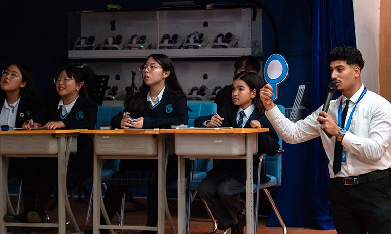 Abdul stands with his class at desks beside him on a stage
