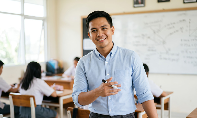 A young male teacher stood in a classroom holding a whiteboard marker