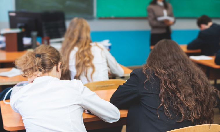 A class of secondary school students at their desks watching a teacher talking at the front of the classroom
