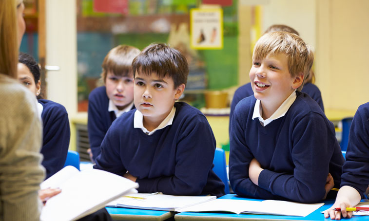 Primary schoolchildren sat at desks as a teacher talks to them from the front of the classroom
