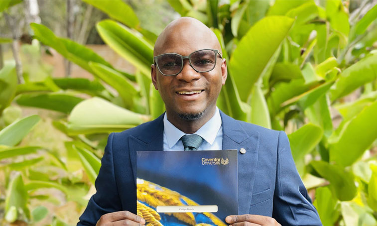 Chilipo Gunda stood outside against a green jungle plants background, holding his MA in Educational Leadership certificate from Coventry University