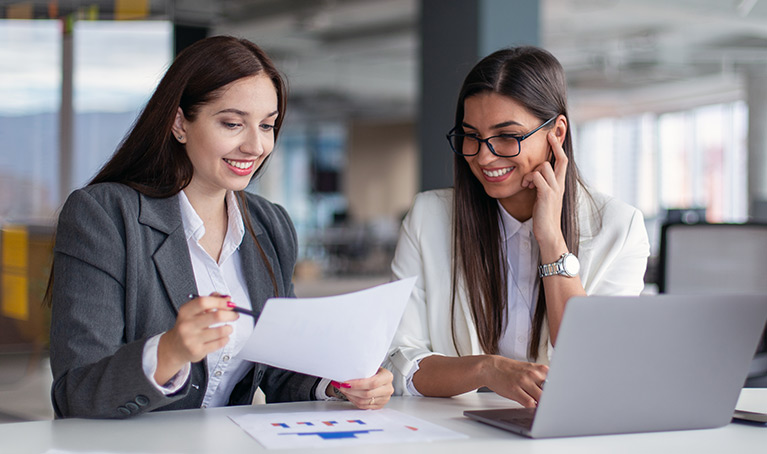 Two women in business wear looking over some paperwork at a table with a laptop open in front of them.