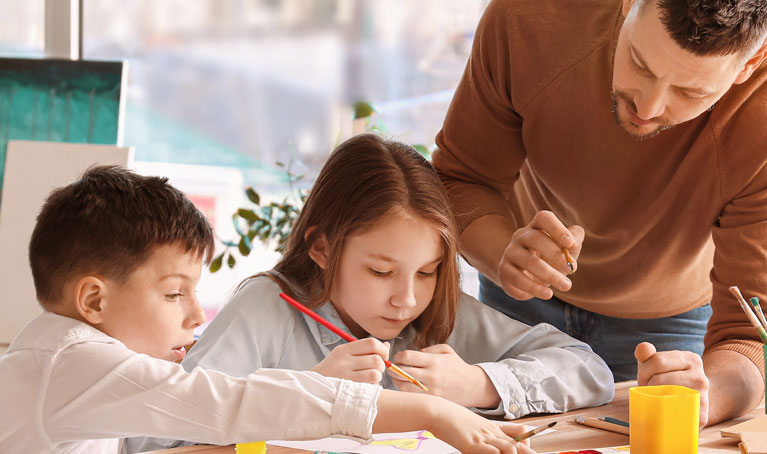A male teaching assistant works with two children at a desk.