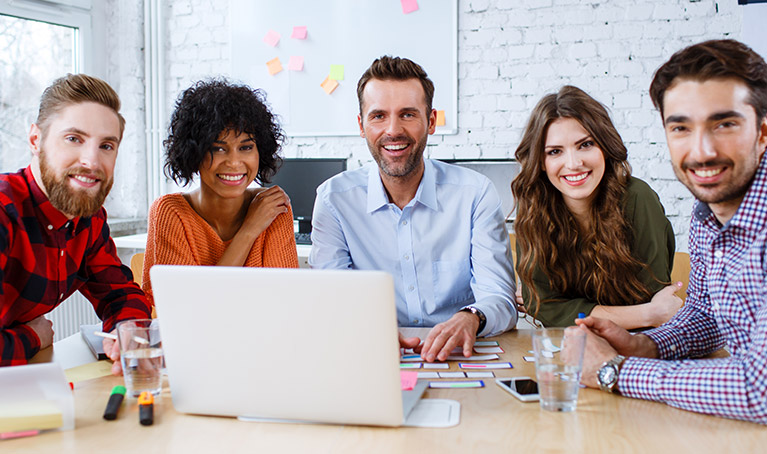 A group of people of various ages sat at a table with a laptop in front of them and smiling at the camera.
