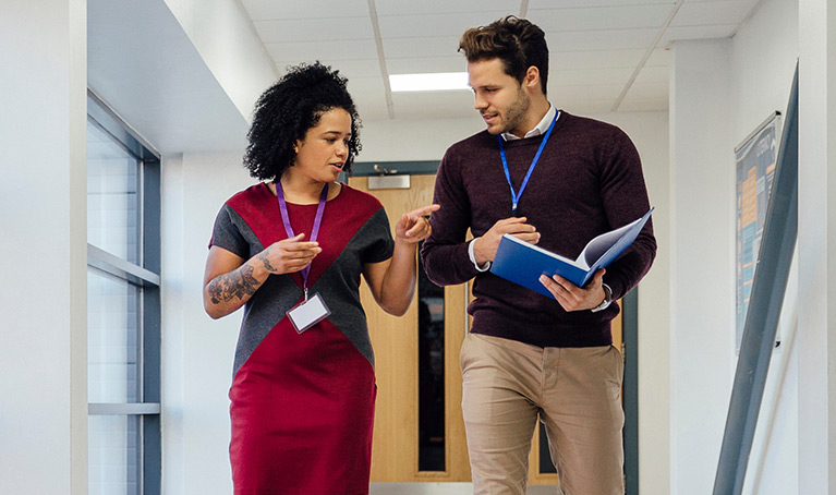 Two teachers walking in a hallway, one carrying an A4 notebook.