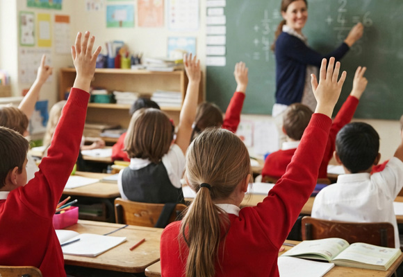 Primary school students in red uniforms sitting at wooden desks inside a bright classroom, raising their hands to answer questions for their teacher
