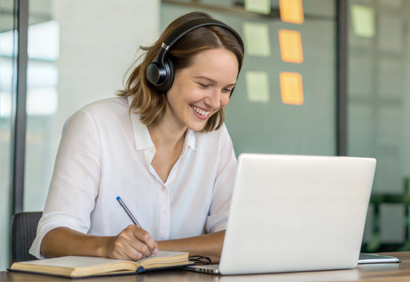 A woman wearing headphones as she takes notes while looking at a laptop