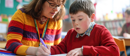 A teacher in a colourful jumper assisting a boy with Down syndrome in a red school uniform in a classroom