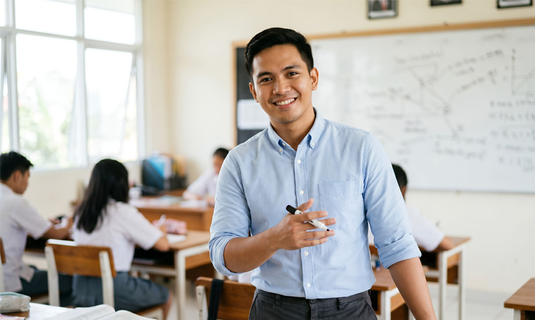 A young male teacher stood in a classroom holding a whiteboard marker