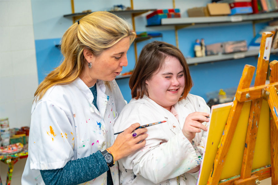 An art teacher helping a young pupil with Down syndrome paint on an easel.
