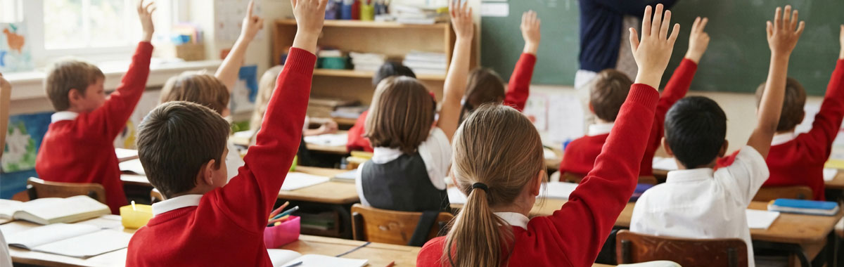 Primary school students in red uniforms sitting at wooden desks inside a bright classroom, raising their hands to answer questions for their teacher