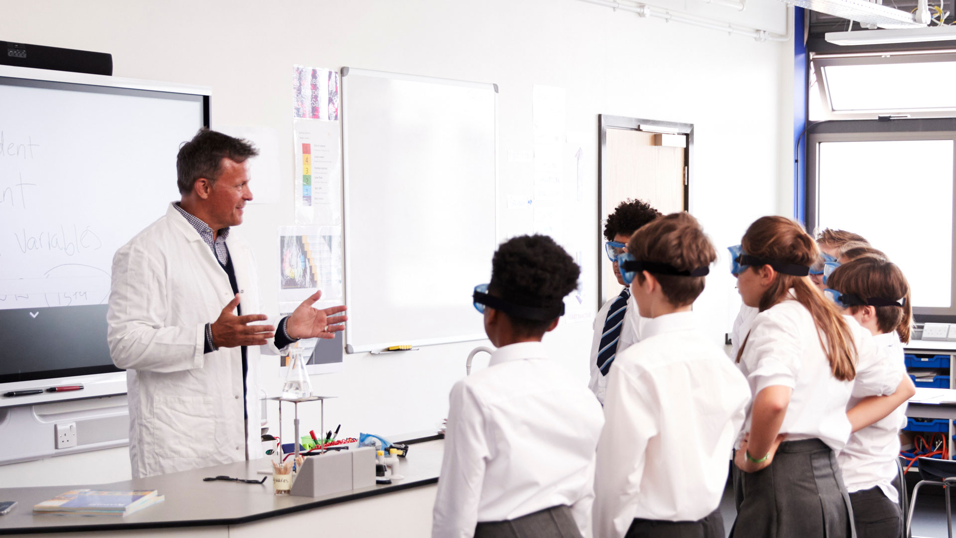 A teacher in a lab coat with a flask on the desk in front of him, talking to a group of students wearing safety glasses.