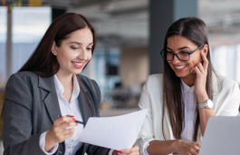 Two women in professional workwear at a desk with a laptop, looking over some papers and smiling.