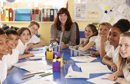 A teacher and students in the classroom sat around a large table, with papers in front of them, all smiling at the camera.