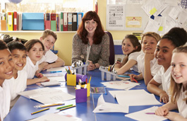 A teacher and students in the classroom sat around a large table, with papers in front of them, all smiling at the camera.