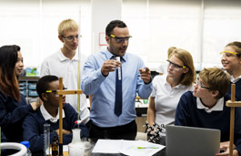 A teacher in a chemistry lab holding a test tube up in front of students wearing safety glasses.