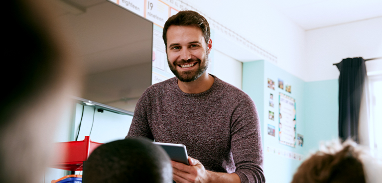 Male teacher stood at the front of a class holding a book