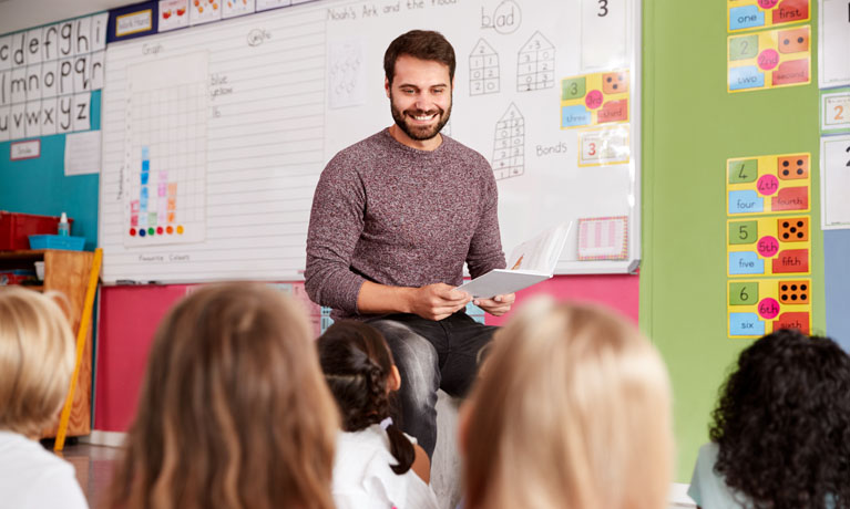 A smiling teacher in front of a white board, pupils seen from behind in foreground