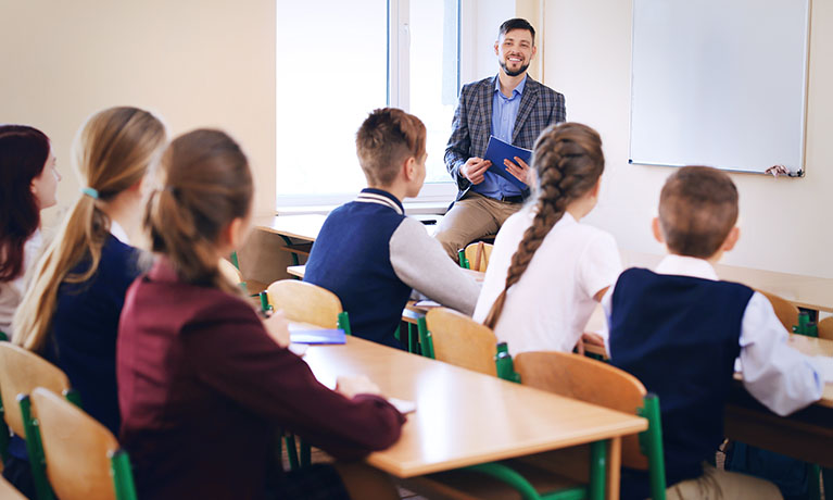 Teacher at front of class talking, pupils sitting at desks.