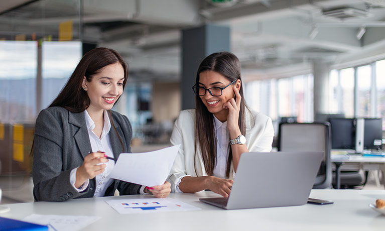Two women in business wear looking over some paperwork at a table with a laptop open in front of them.