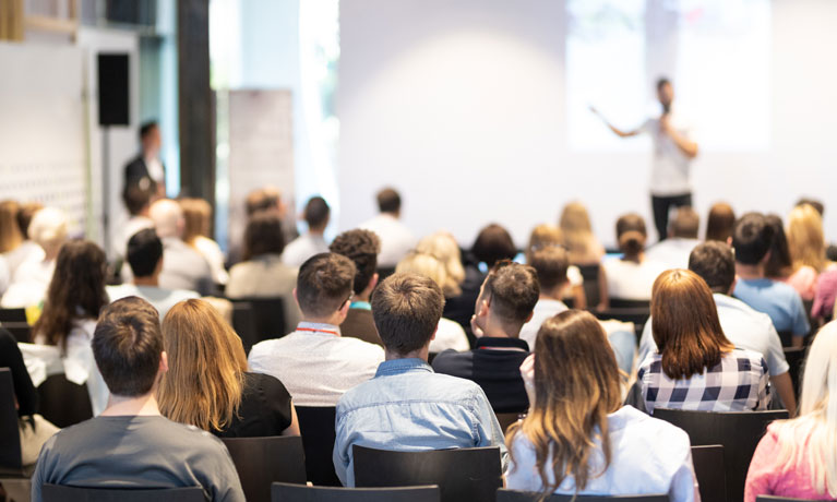 A group of people sat down listening to a speaker presenting at the front of the room