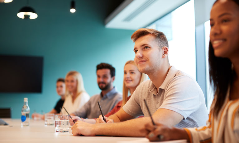 Group of young females and males sitting in an office looking towards a screen