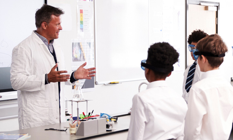 A teacher in a lab coat talking to students in safety goggles.