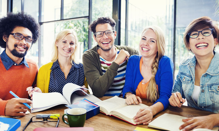 Five mature students in colourful clothing sat together at a table with notebooks, smiling at the camera.