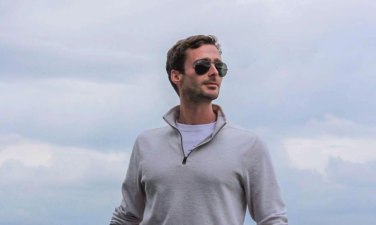 An image of Aaron, wearing a grey jumper and sunglasses, smiling into the distance.