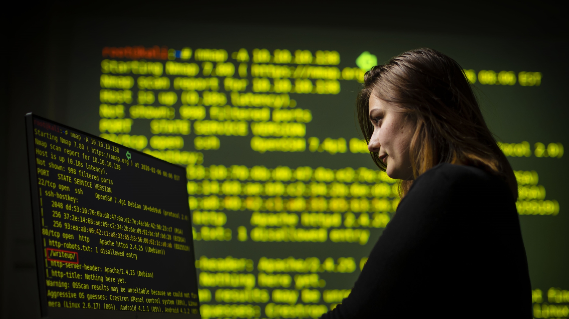 Woman looking down at computer screen with large screen in background showing lines of text