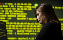 Woman looking down at computer screen with large screen in background showing lines of text